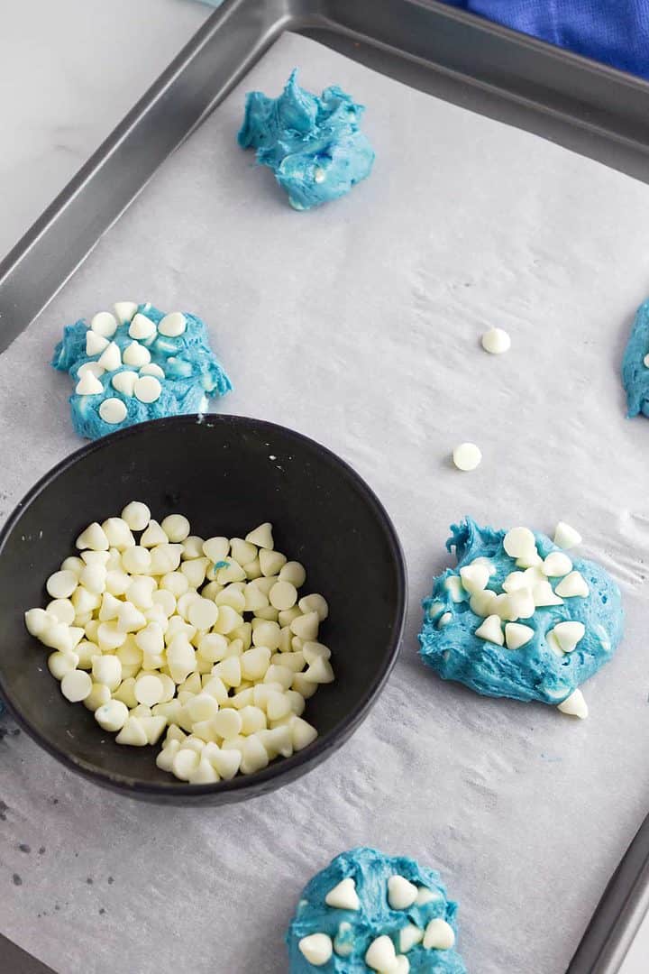 Blue velvet cookie dough with white chocolate chips on a parchment-lined tray; black bowl of extra chips beside it.