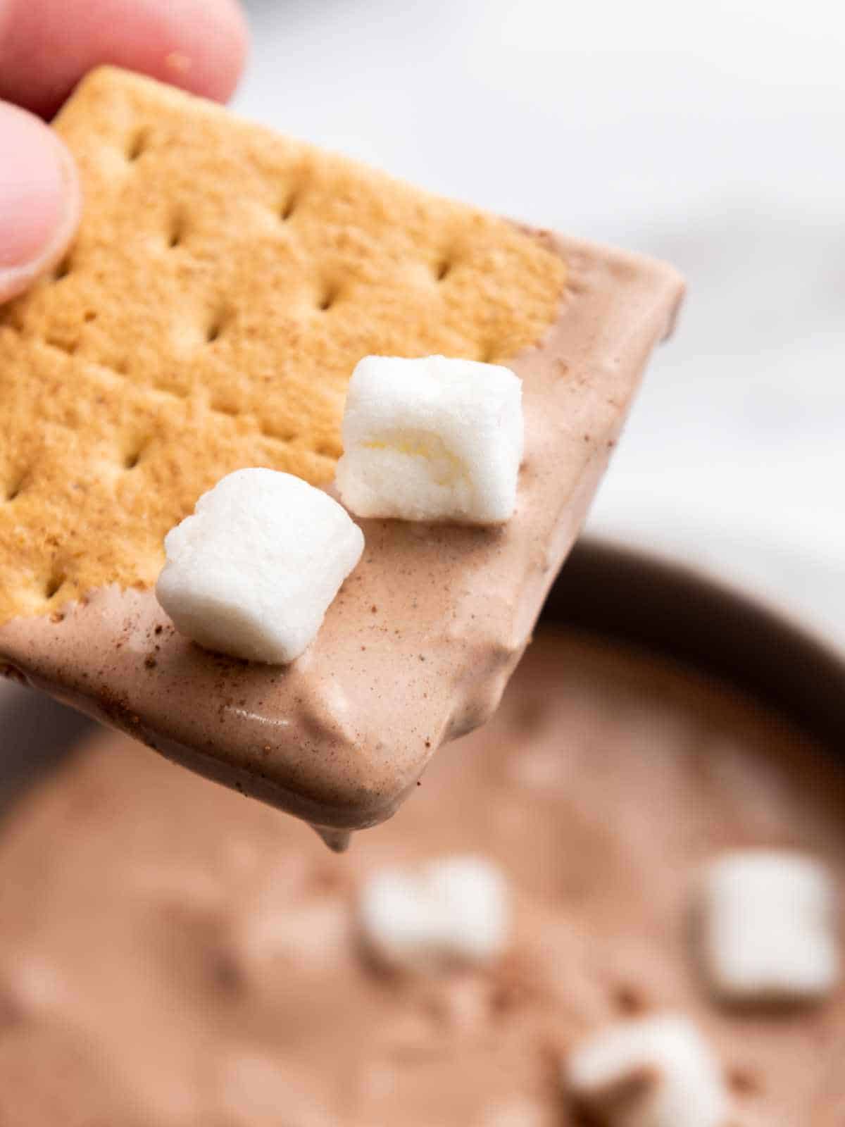 Graham cracker with hot cocoa dip and two mini marshmallows with dip in background.