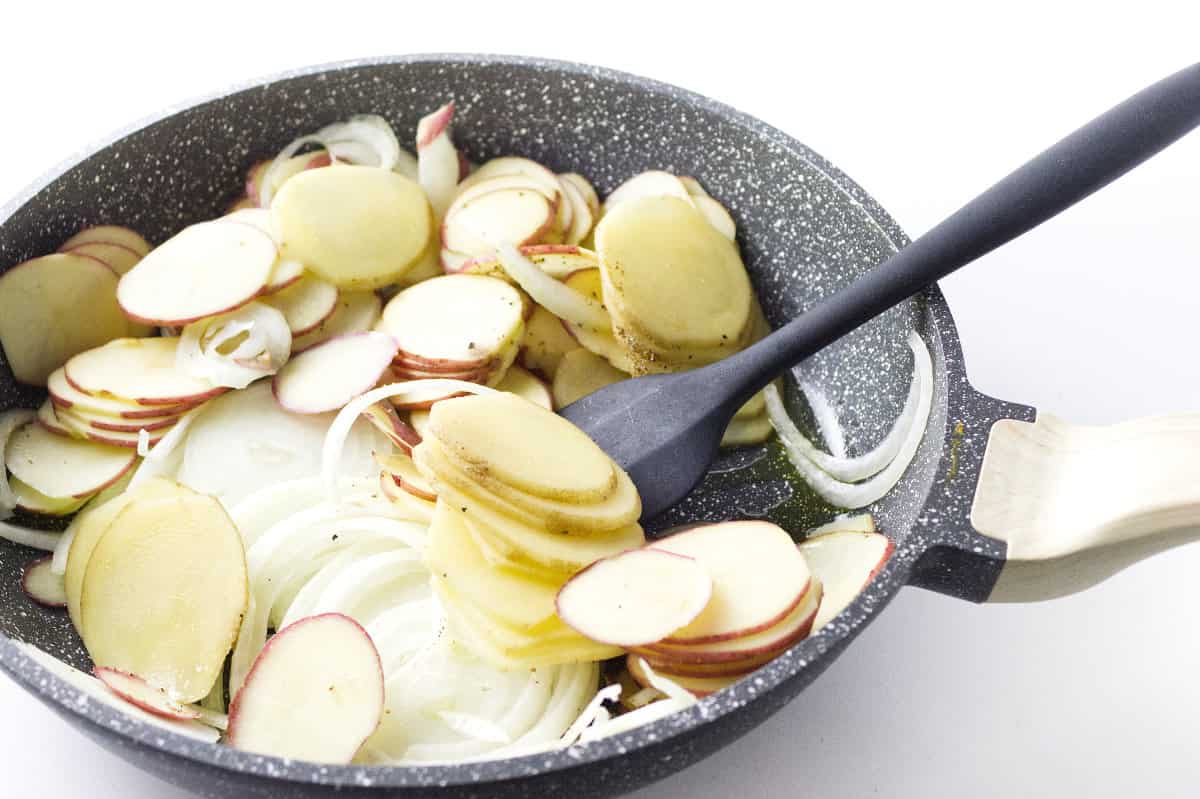 skillet with spatula stirring sliced onions, sliced potatoes, and seasoninggs.