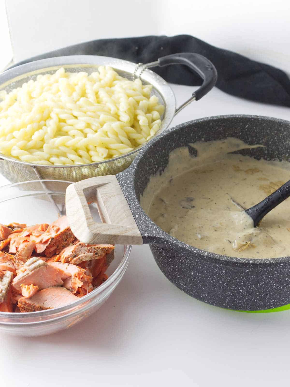 colander of drained pasta, bowl of salmon chunks, and pot of mushroom alfredo sauce.
