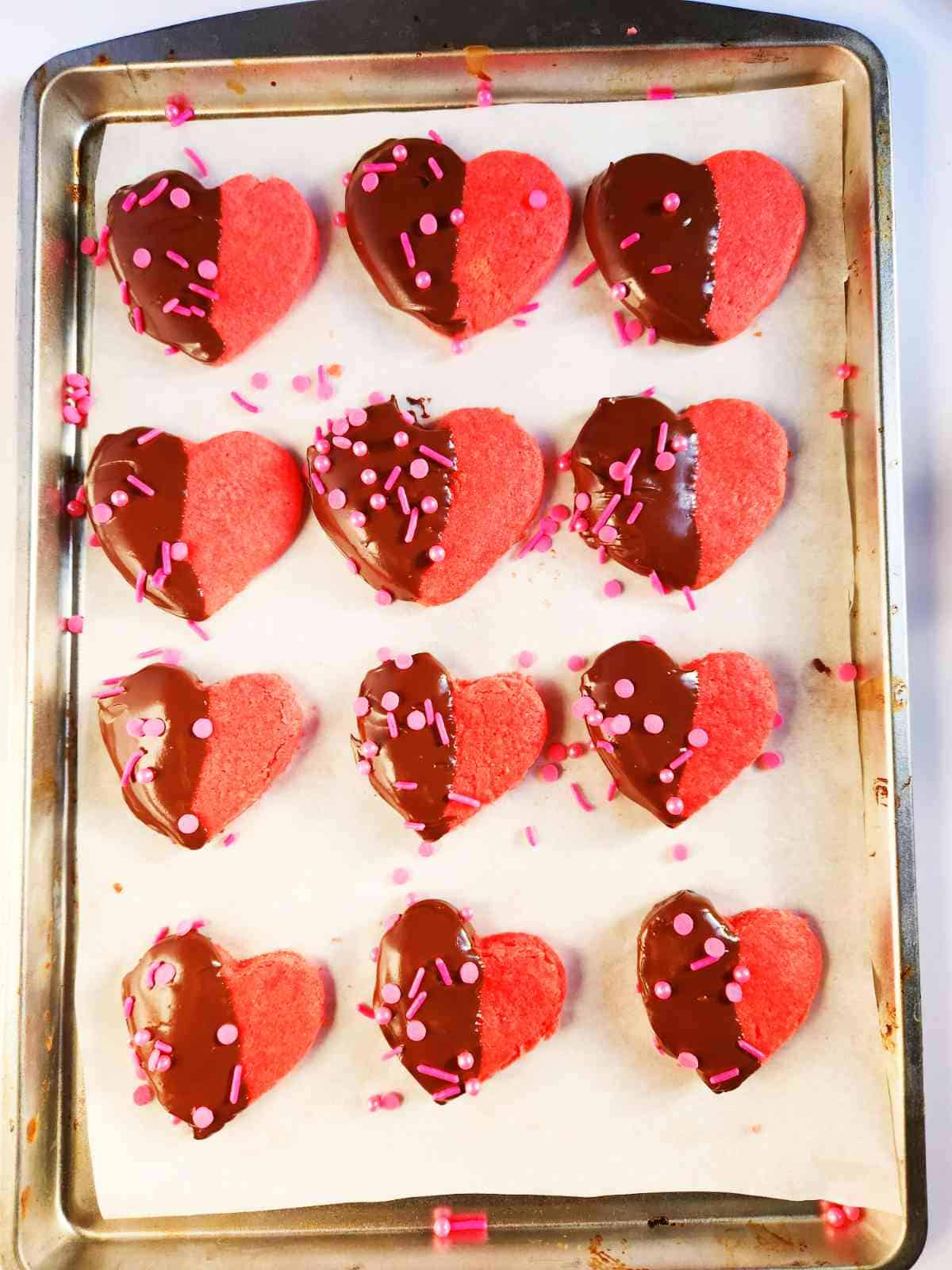 sprinkles on chocolate dipped pink heart cookies on a parchment lined baking sheet.