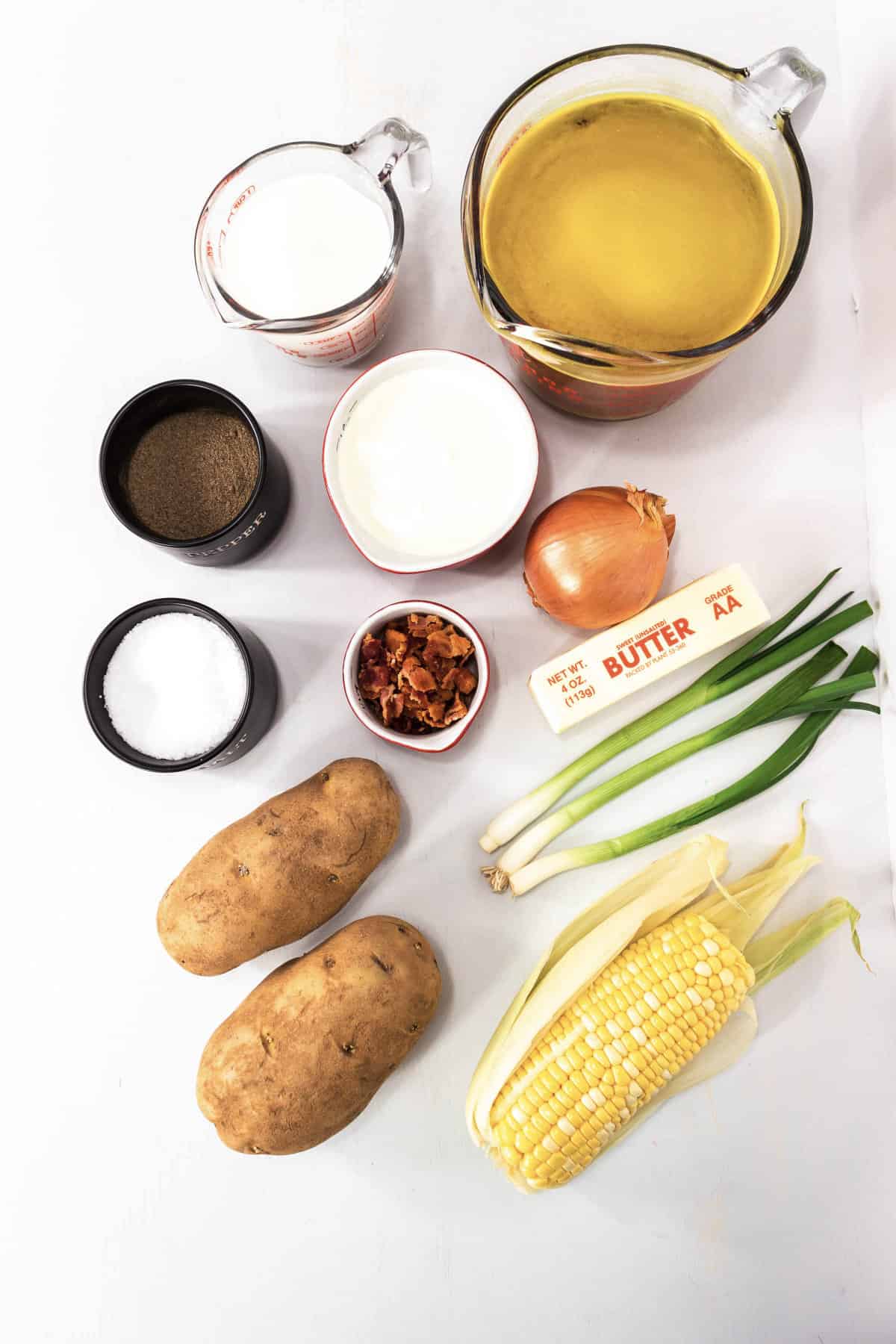 ingredients on a white counter to make soup with potatoes, corn, bacon, onion, butter, milk, and broth.