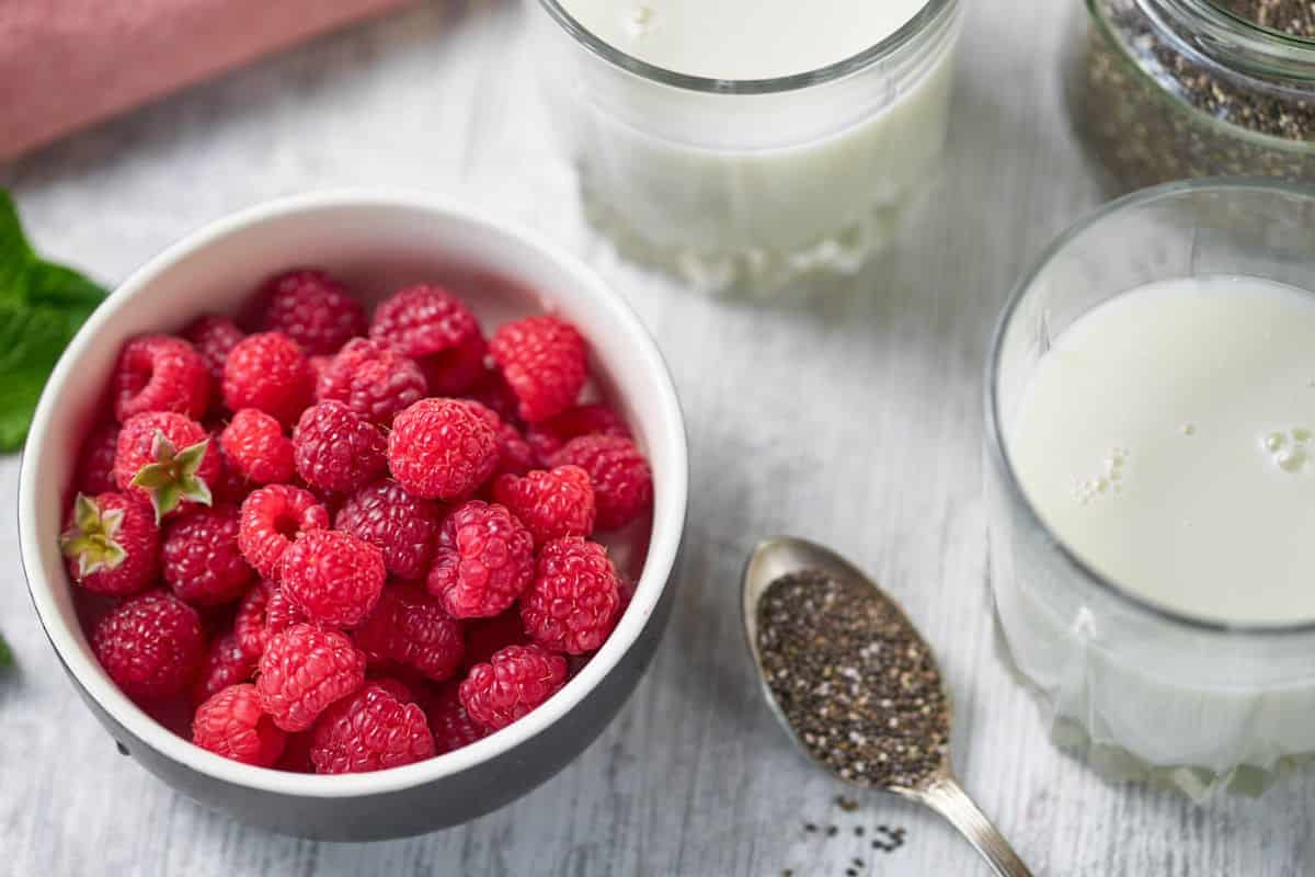 two glasses of milk, a spoonful of seeds, and a bowl of fresh raspberries.