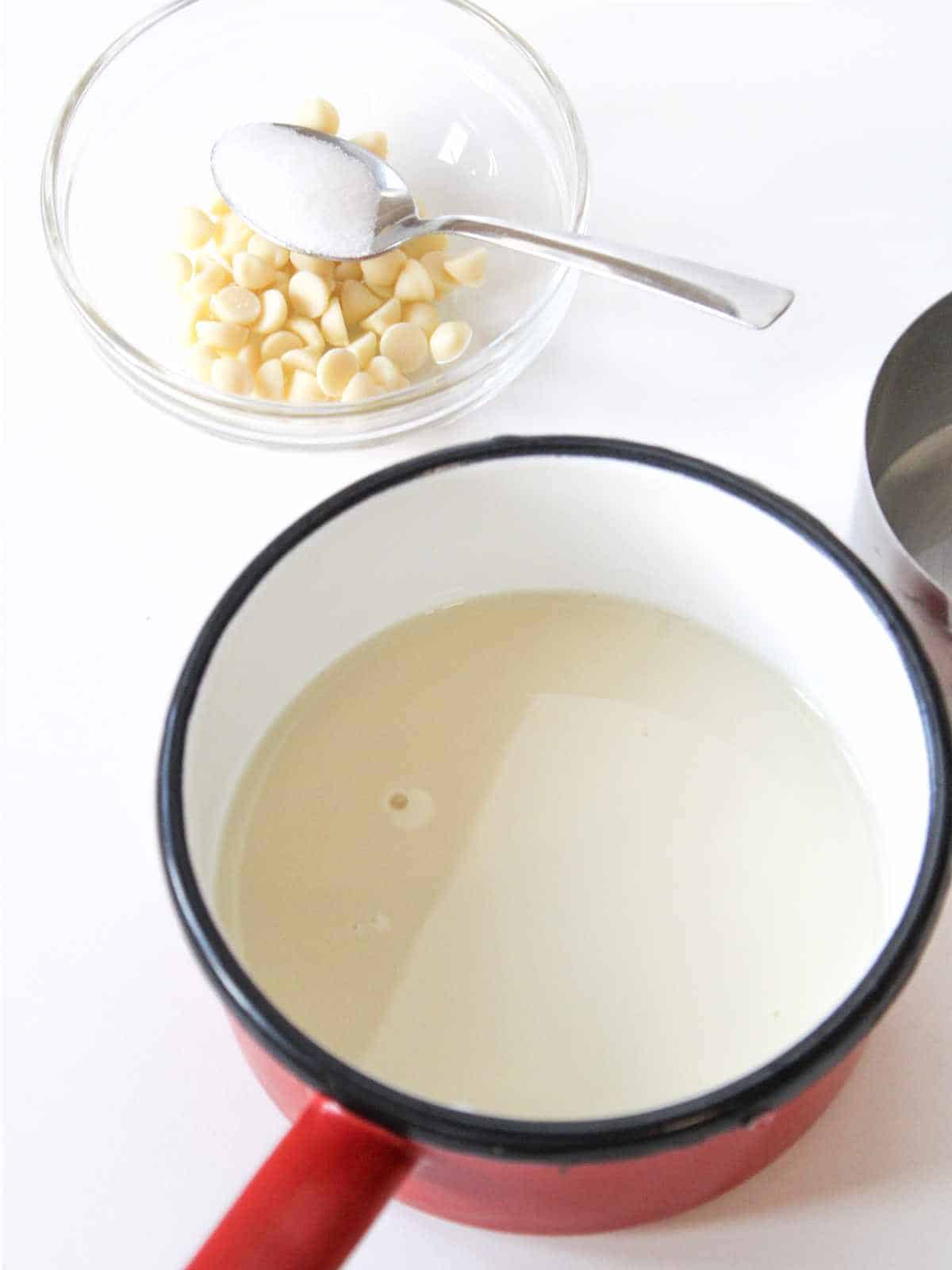 Hot milk in a sauce pan with white chocolate chips in a bowl nearby.