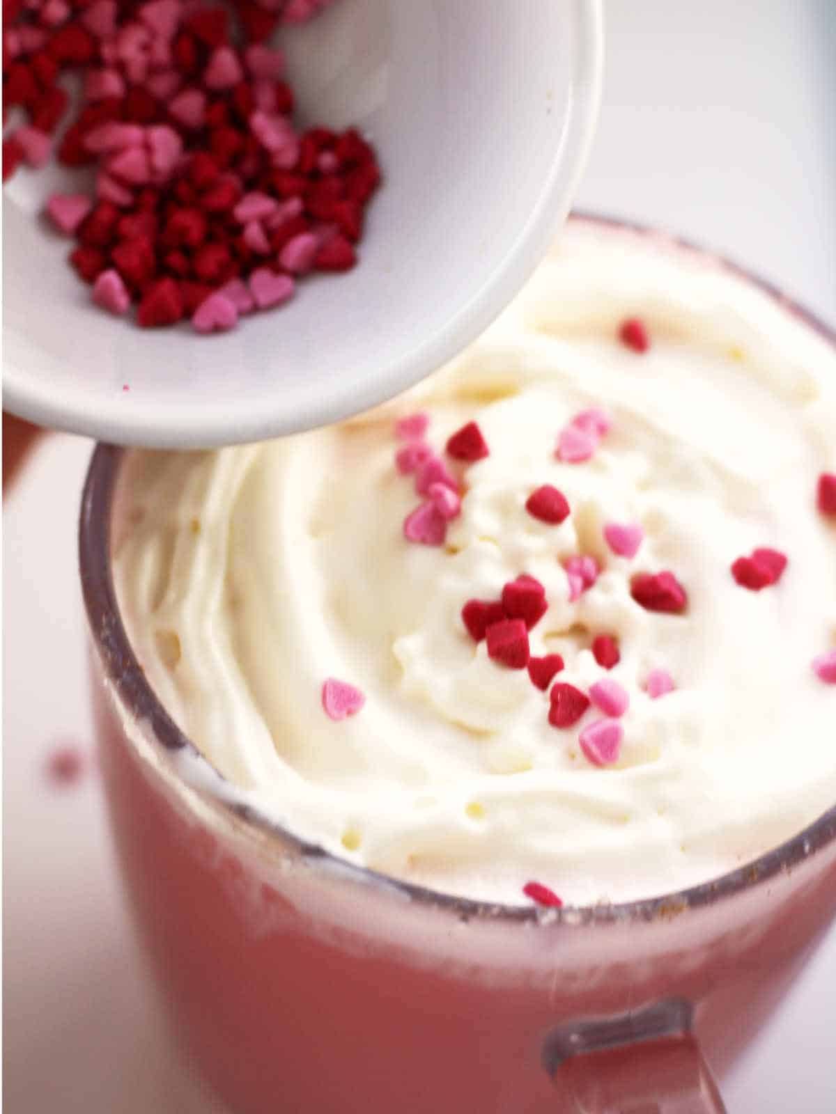 Sprinkling red and pink heart sprinkle on top of whip cream topped hot cocoa.