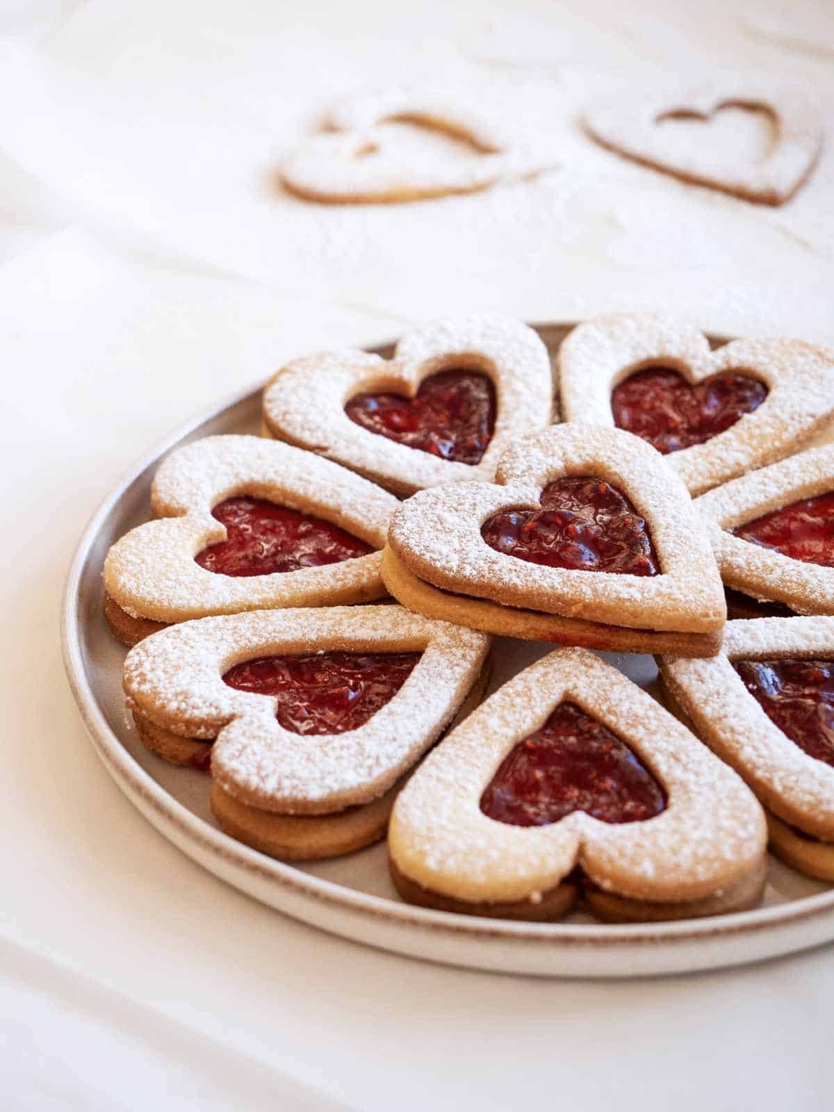 Plate of Raspberry Linzer cookies with sugar dusted cutout tops in the background.