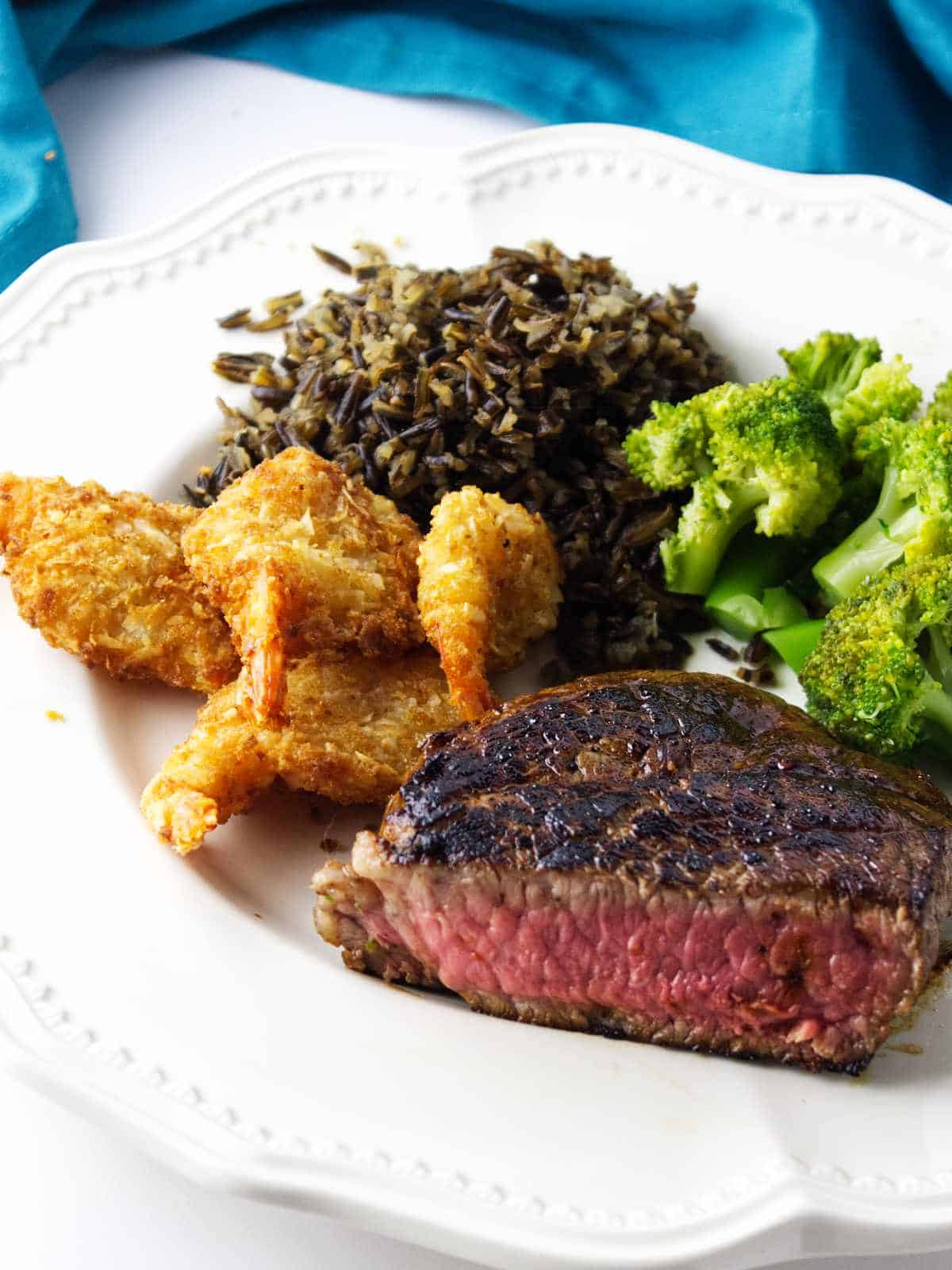 serving of breaded shrimp, steak, broccoli, and wild rice on a white plate.
