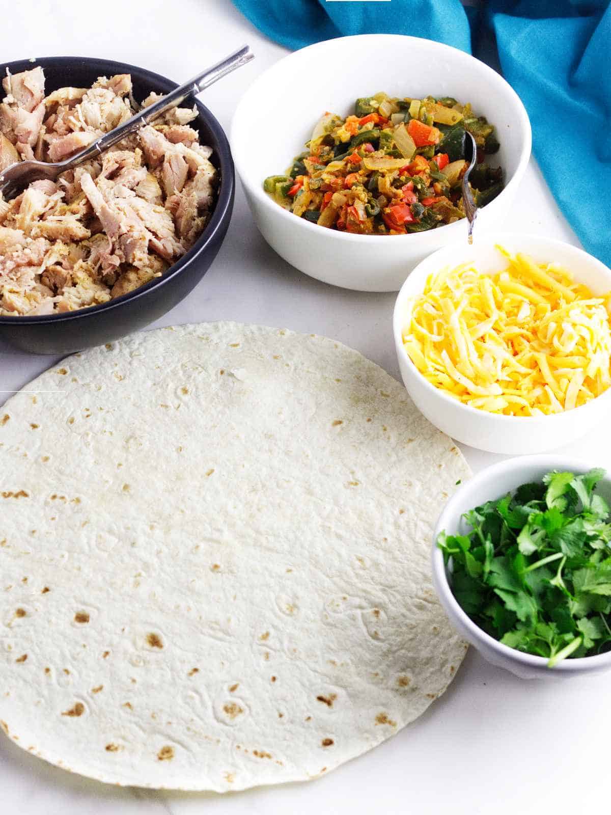 flour tortillas with bowls of shredded chicken, shredded cheese, sauteed peppers and onions, and chopped cilantro.