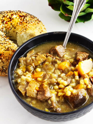 a bowl of beef and barley soup with a bagel side.