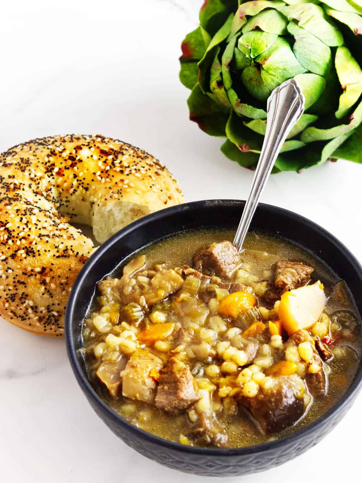 a bowl of beef  barley soup with a bagel side.