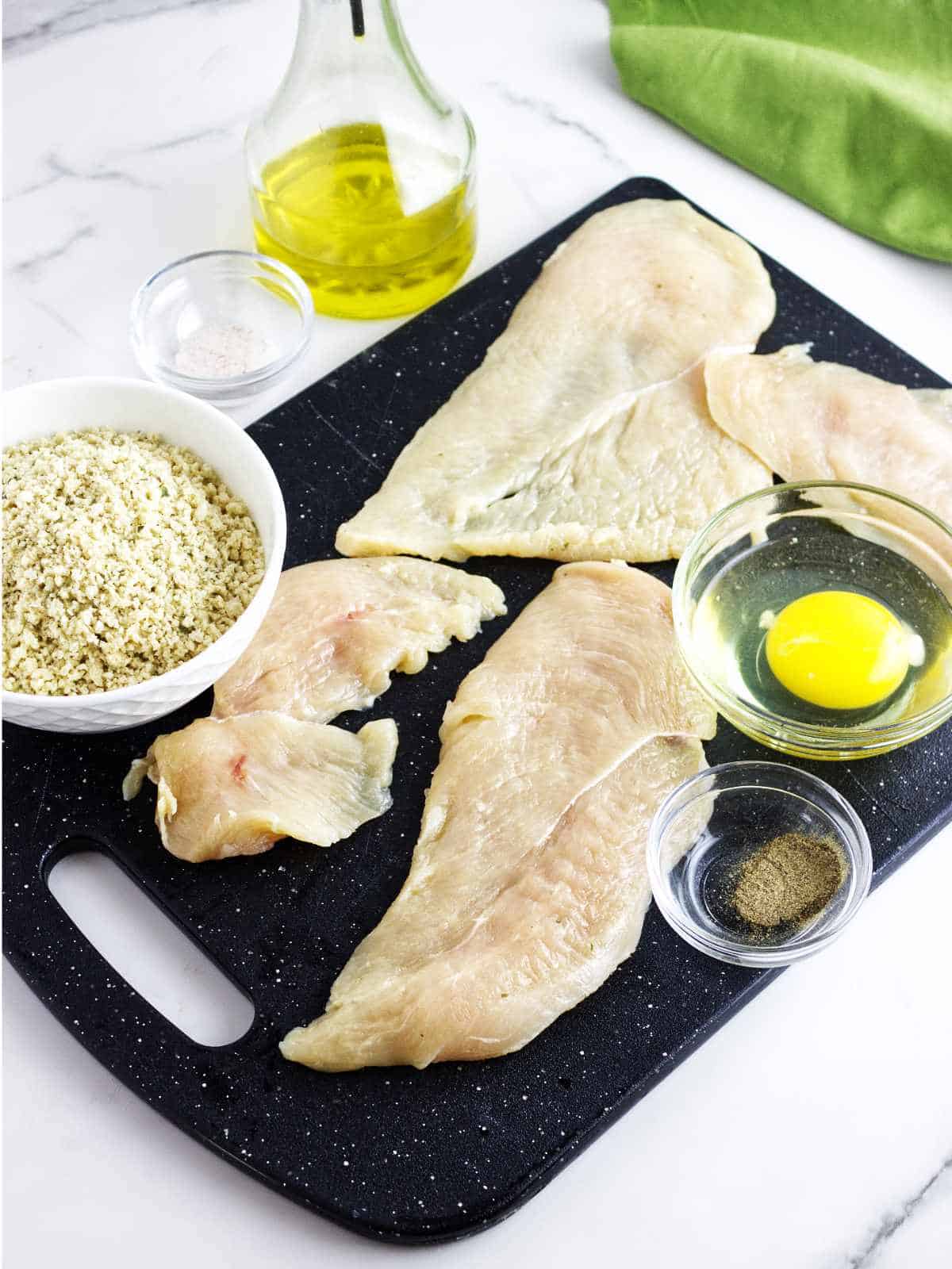cutting board with thin sliced chicken breast, panko bread crumbs, egg, and seasoning.