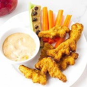 Child's lunch plate of air fried dino nuggets, carrot sticks, raisins on a peanut butter and celery stick, and dip on a white plate with a red apple nearby.
