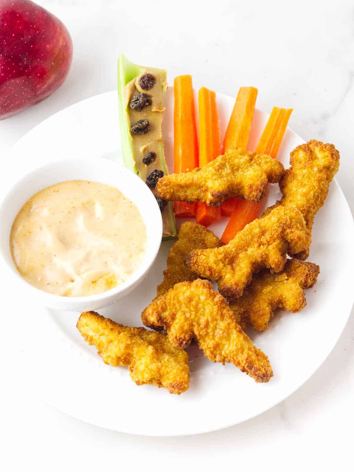 Child's lunch plate of air fried dino nuggets, carrot sticks, raisins on a peanut butter and celery stick, and dip on a white plate with a red apple nearby.