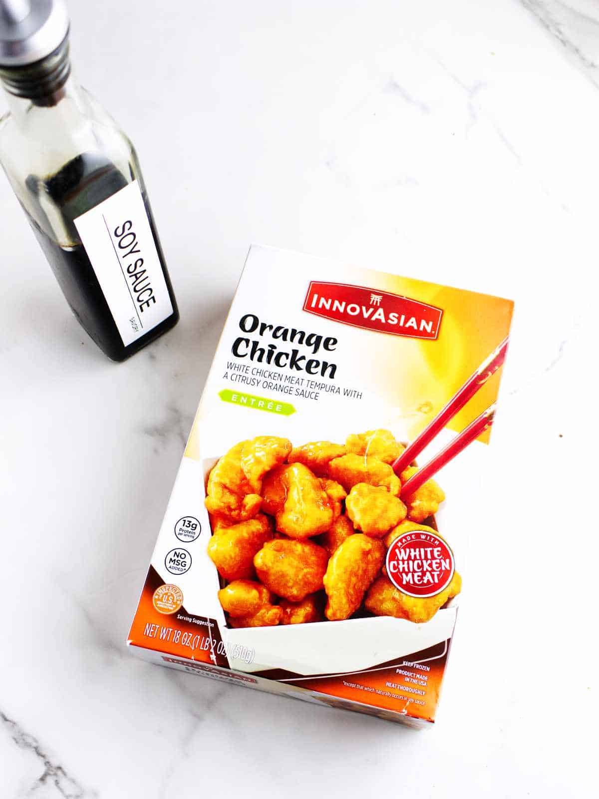 package of frozen prepared Innovasian white chicken tempura with orange sauce, and a bottle of savory soy sauce on a white background.