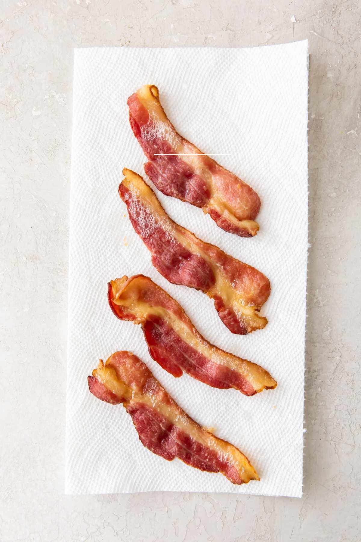 Four cooked strips of air fryer bacon are laid out on a white paper towel, which is placed on a light-colored, textured surface.