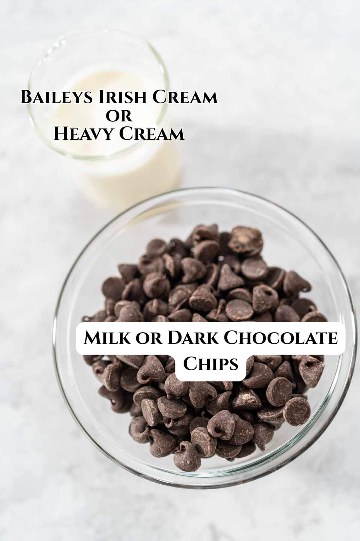 A glass bowl filled with chocolate chips for baileys Irish Cream Ganache sits next to a small glass of milk on a light gray surface.