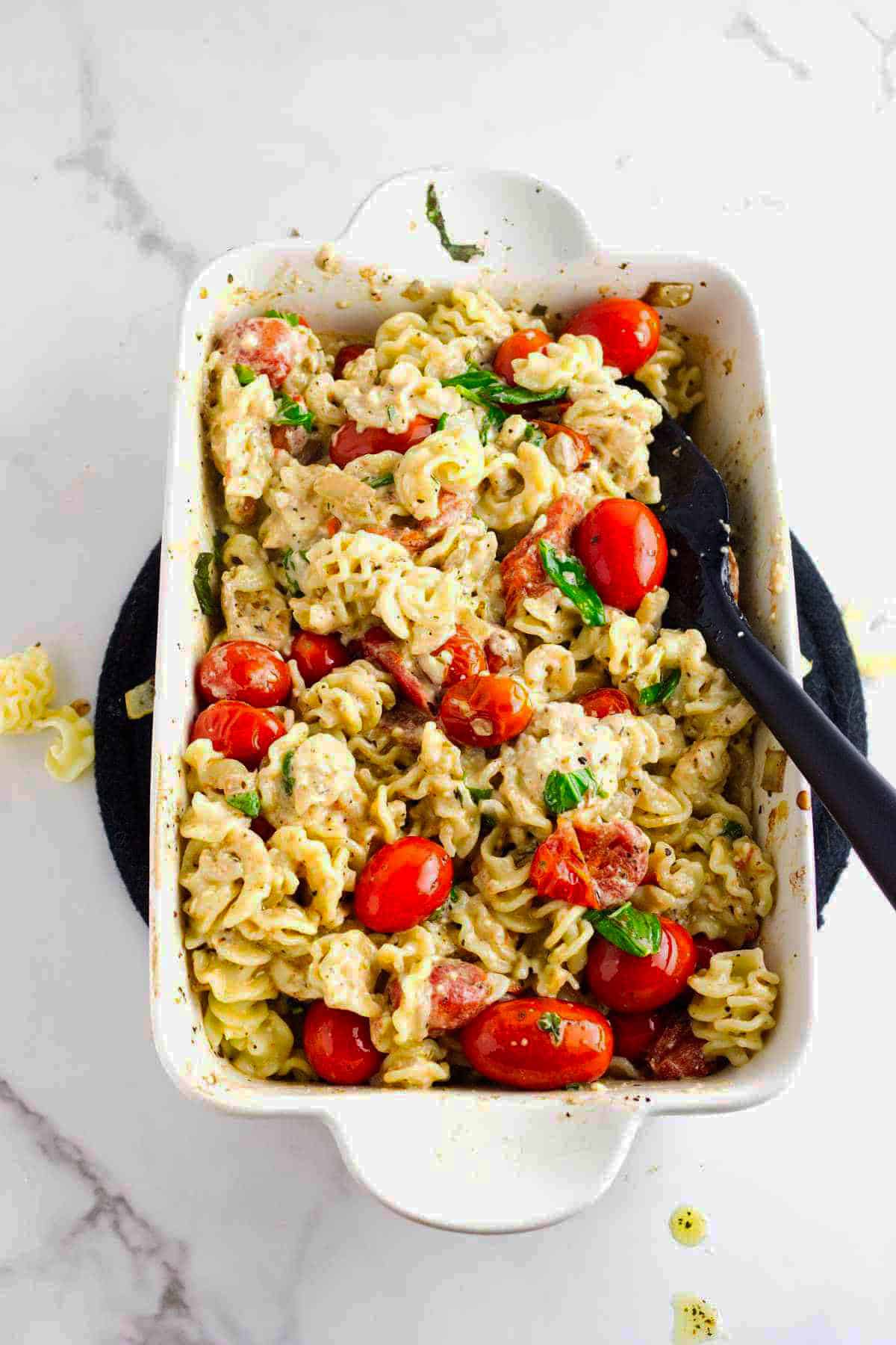 Baked feta cheese and tomato pasta with rotini in a creamy sauce fills a white dish. A black serving spoon rests in the pasta, which is topped with fresh herbs. The dish sits on a dark mat atop a white surface.