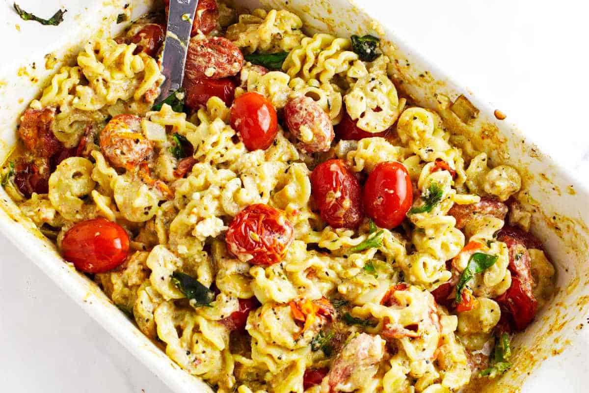 A baking dish filled with feta cheese and tomato pasta, spinach, and a creamy sauce, mixed together and baked until golden, with a serving spoon in the dish.