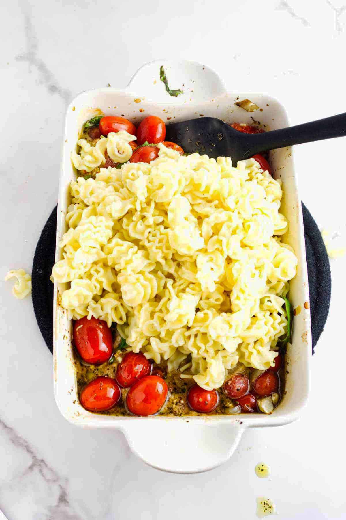 A white baking dish filled with curly feta cheese and tomato pasta, cherry tomatoes, and seasonings sits on a dark trivet. A black spoon rests inside the dish, with some sauce visible at the bottom.