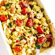 Baked pasta dish with melted cheese, cherry tomatoes reminiscent of feta cheese and tomato pasta, sausage slices, spinach, and spiral-shaped noodles in a white casserole dish with a serving spoon.