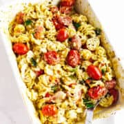 Baked feta cheese and tomato pasta in a white dish with melted cheese, roasted cherry tomatoes, spiral noodles, spinach, and herbs, with a serving spoon placed inside.