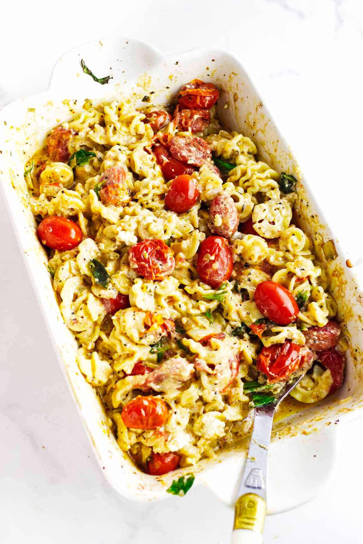 Baked feta cheese and tomato pasta in a white dish with melted cheese, roasted cherry tomatoes, spiral noodles, spinach, and herbs, with a serving spoon placed inside.
