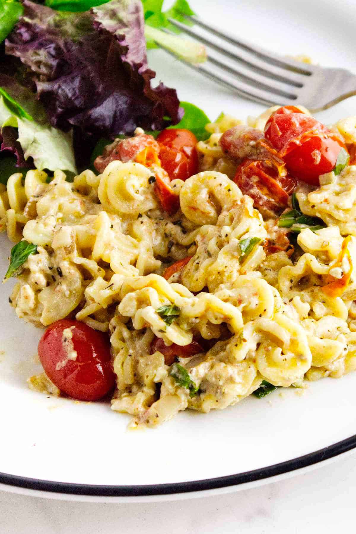 A plate of creamy feta cheese and tomato pasta with cherry tomatoes, fresh basil, and visible herbs, served with a side of mixed leafy greens. A fork rests behind the food on the white plate.