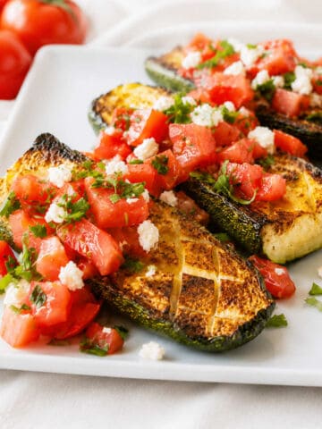 Baked zucchini half on a white plate, topped with tomatoes and crumbled feta with fresh parsley.