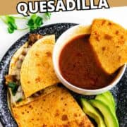 A plate with two folded birria quesadillas, sliced avocado, and a bowl of red dipping sauce.