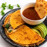 Birria quesadilla dipped into a bowl of rich consome with melted cheese and shredded beef visible.
