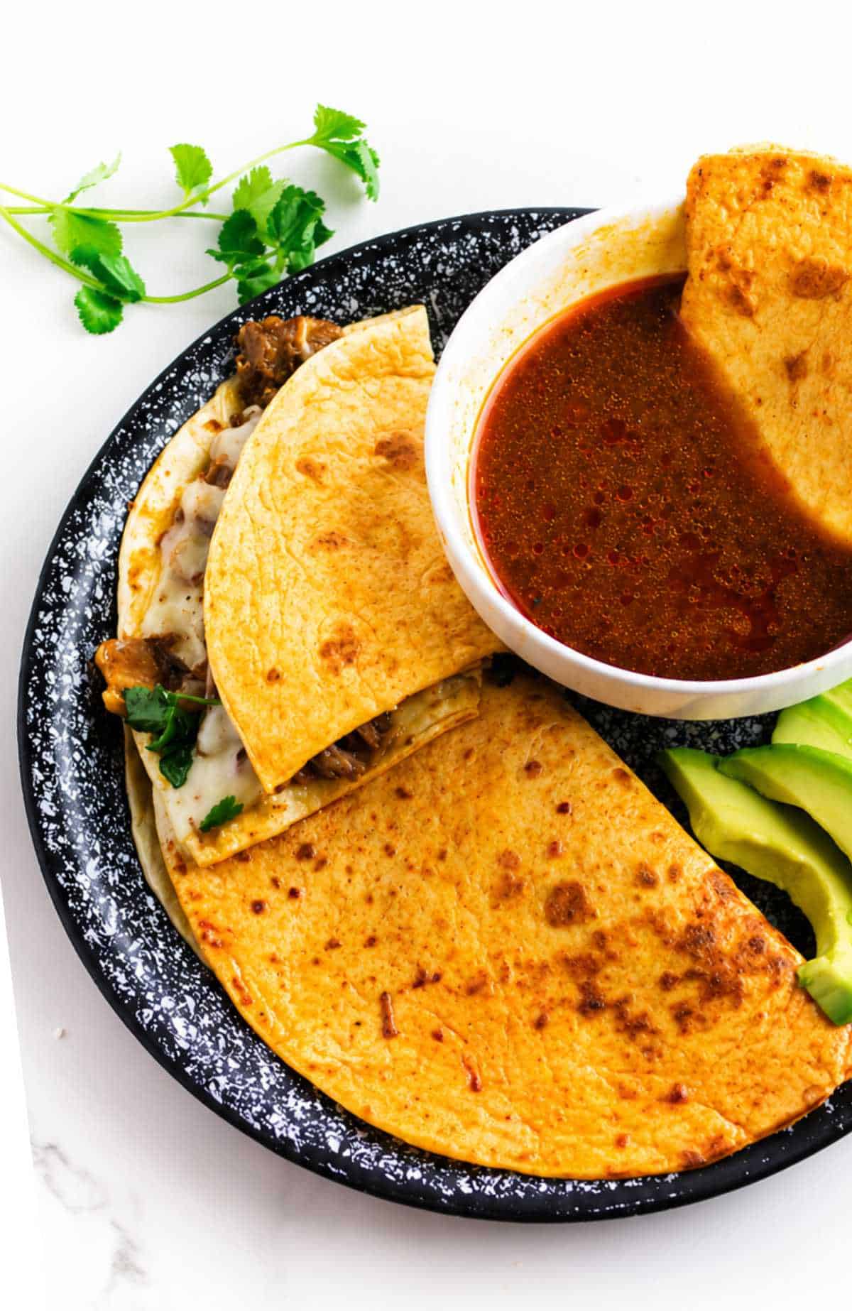 Crispy birria quesadillas stacked on a plate with a bowl of consome on the side.