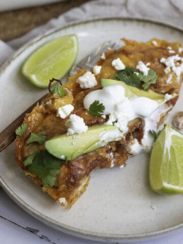 A plate with a serving of cheesy enchiladas topped with avocado slices, sour cream, cilantro, and crumbled cheese, garnished with lime wedges. A fork is resting on the plate with a bite of food.
