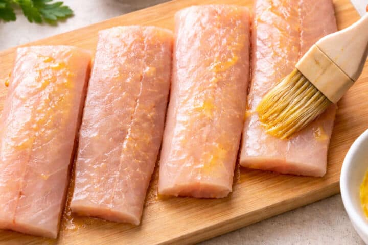 Two raw fish fillets on a white plate being brushed with oil, perfect for making blackened mahi mahi, sit next to a bowl of spice mix and a bottle of olive oil on a white marble surface.