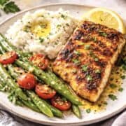 Two seasoned and baked fish fillets are served on a white plate, garnished with lemon wedges. The fish has a golden-brown crust from spices.