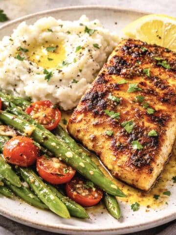 Two seasoned and baked fish fillets are served on a white plate, garnished with lemon wedges. The fish has a golden-brown crust from spices.