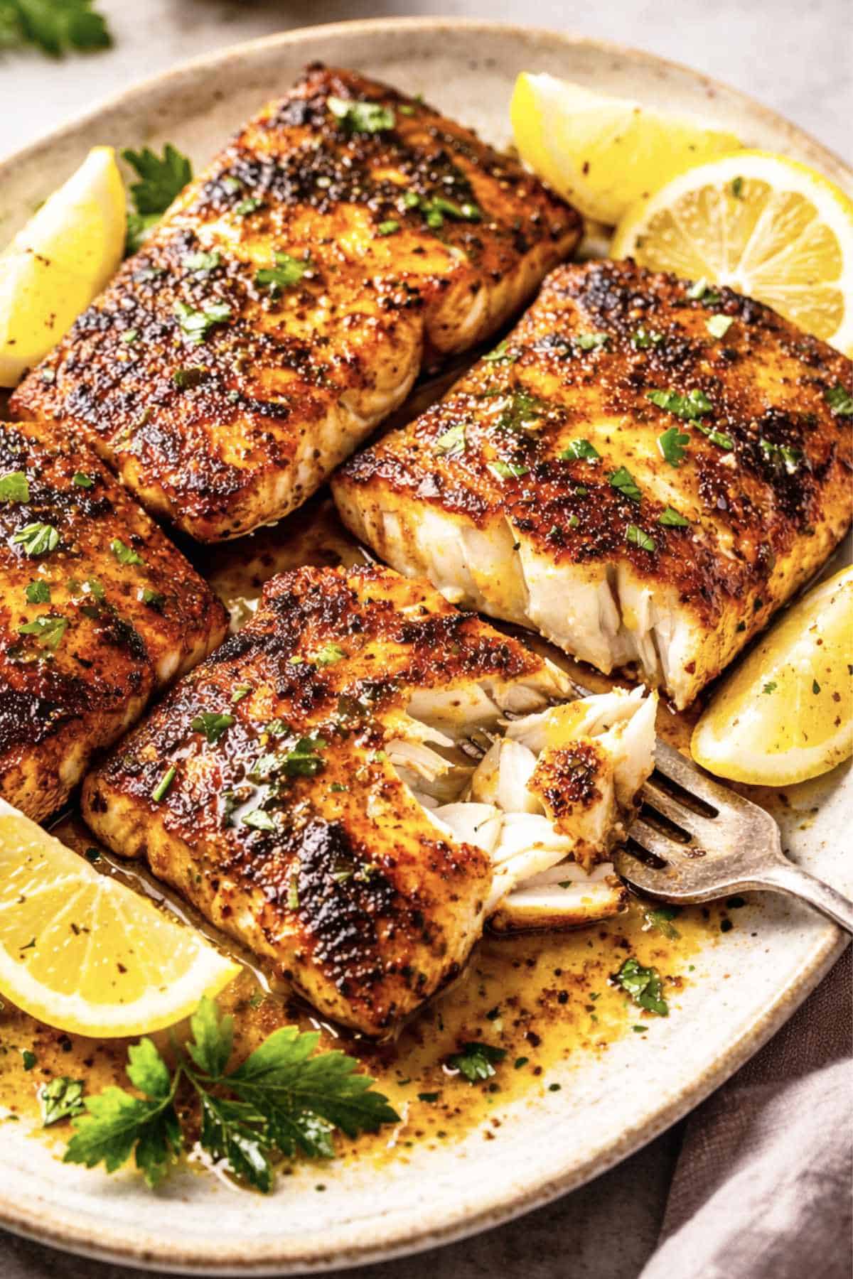 Two seasoned and baked fish fillets—reminiscent of blackened mahi mahi—rest in a white dish, garnished with lemon slices. A bowl of creamy white sauce with herbs and a spoon sits nearby on a white surface.