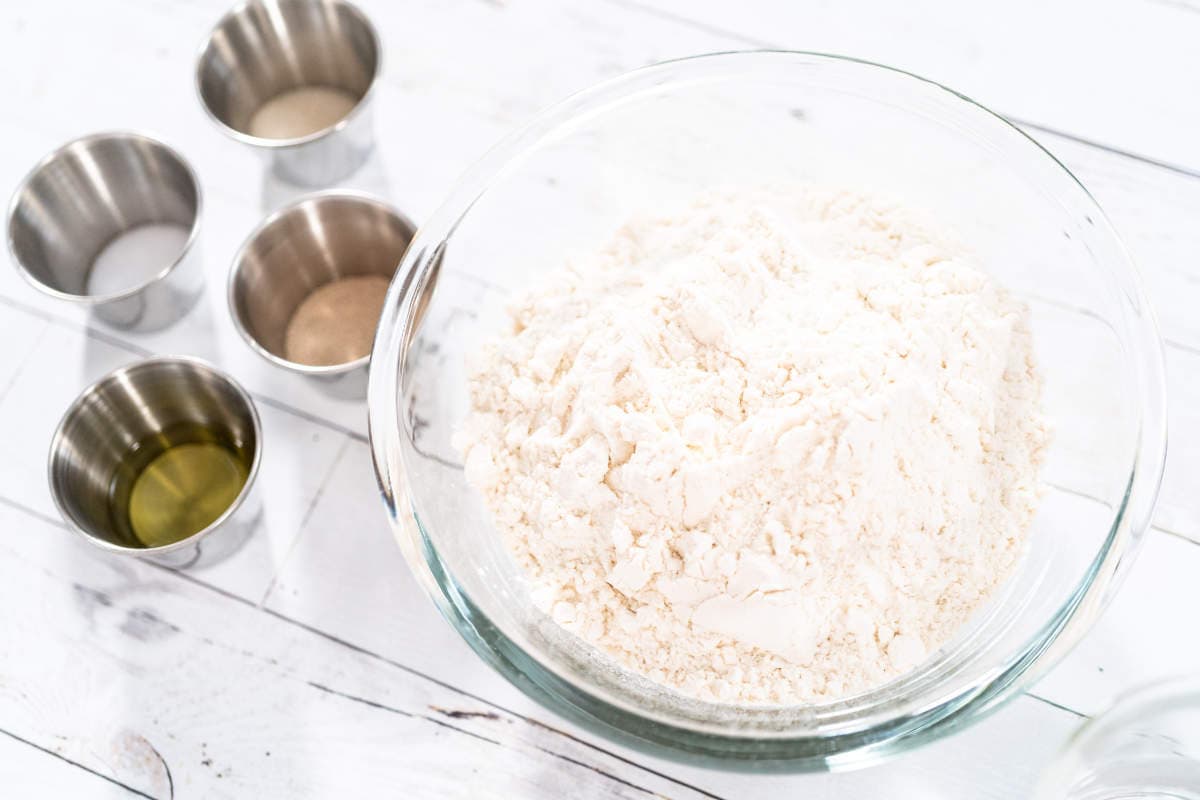 A glass bowl filled with flour sits on a white wooden surface next to four small metal cups containing oil, yeast, salt, and another ingredient.