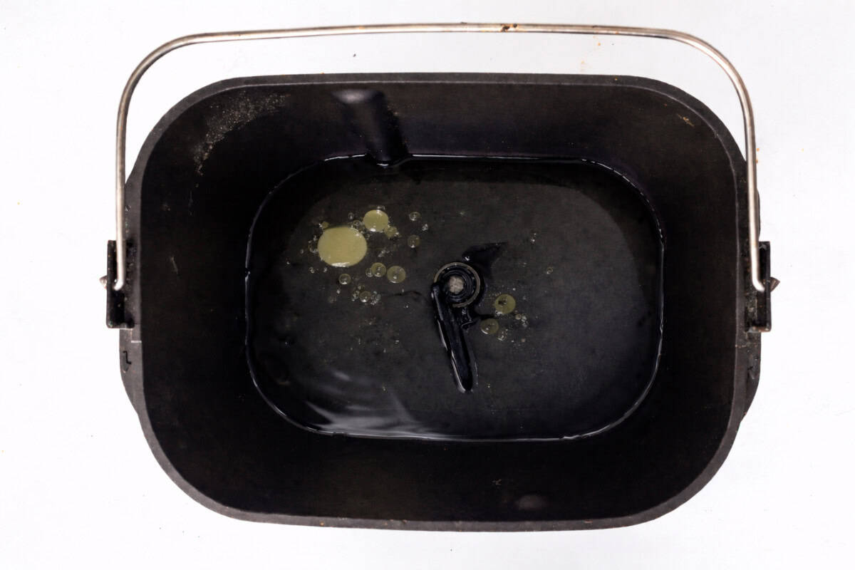 A black bread machine pan with a handle, containing water and patches of oil, viewed from above on a white background.
