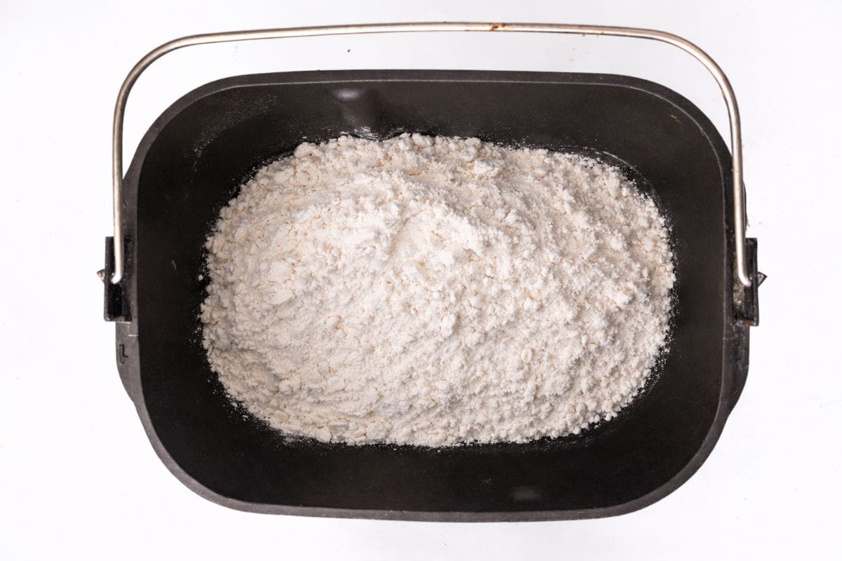 A black bread machine pan filled with white flour, viewed from above, against a plain white background.