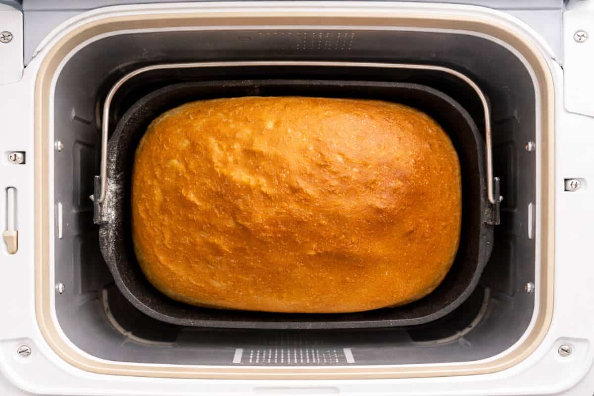 A golden-brown loaf of bread sits inside a bread machine pan, freshly baked with a slightly textured top crust.