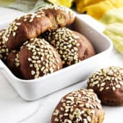 A white dish filled with round, dark brown pumpernickle rolls topped with oats. Two rolls are placed on a white surface next to the dish. A yellow cloth is partially visible in the background.