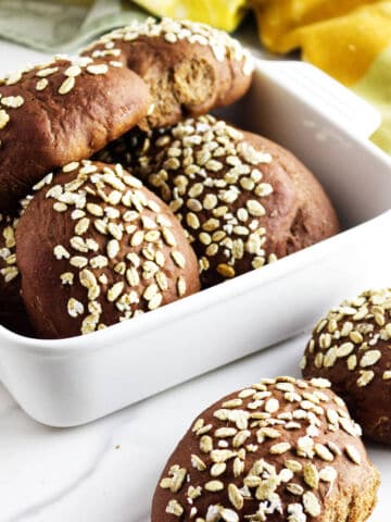 A white dish filled with round, dark brown pumpernickle rolls topped with oats. Two rolls are placed on a white surface next to the dish. A yellow cloth is partially visible in the background.