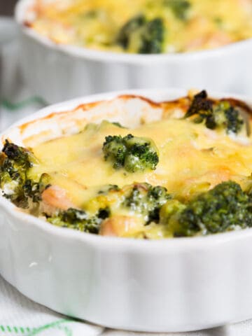 Close-up of a cheesy broccoli and chicken casserole in a white ramekin, with melted cheese on top and slight browning around the edges. A fork and napkin are visible beside the dish.