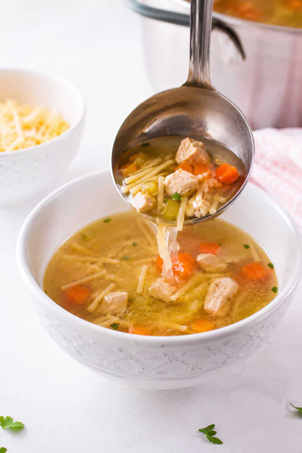 A ladle of chicken fideo soup with chicken, noodles, and vegetables is served into a white bowl; steaming pot behind.
