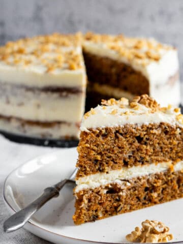 A slice of carrot cake with cream cheese frosting and chopped walnuts sits on a plate with a fork. The rest of the cake, also topped with frosting and walnuts, is in the background with a slice missing.