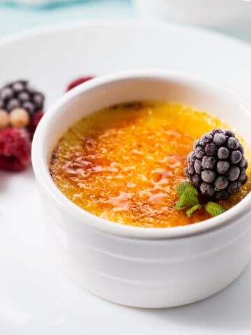 A white ramekin filled with classic crème brûlée, topped with two fresh raspberries and a mint leaf. A spoon is scooping into the creamy dessert, and another ramekin is partially visible in the background.