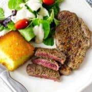 A plate with a grilled Denver steak, sliced to show a pink center, a green salad with grape tomatoes and creamy dressing, and a square piece of cornbread. A fork and knife are beside the plate.