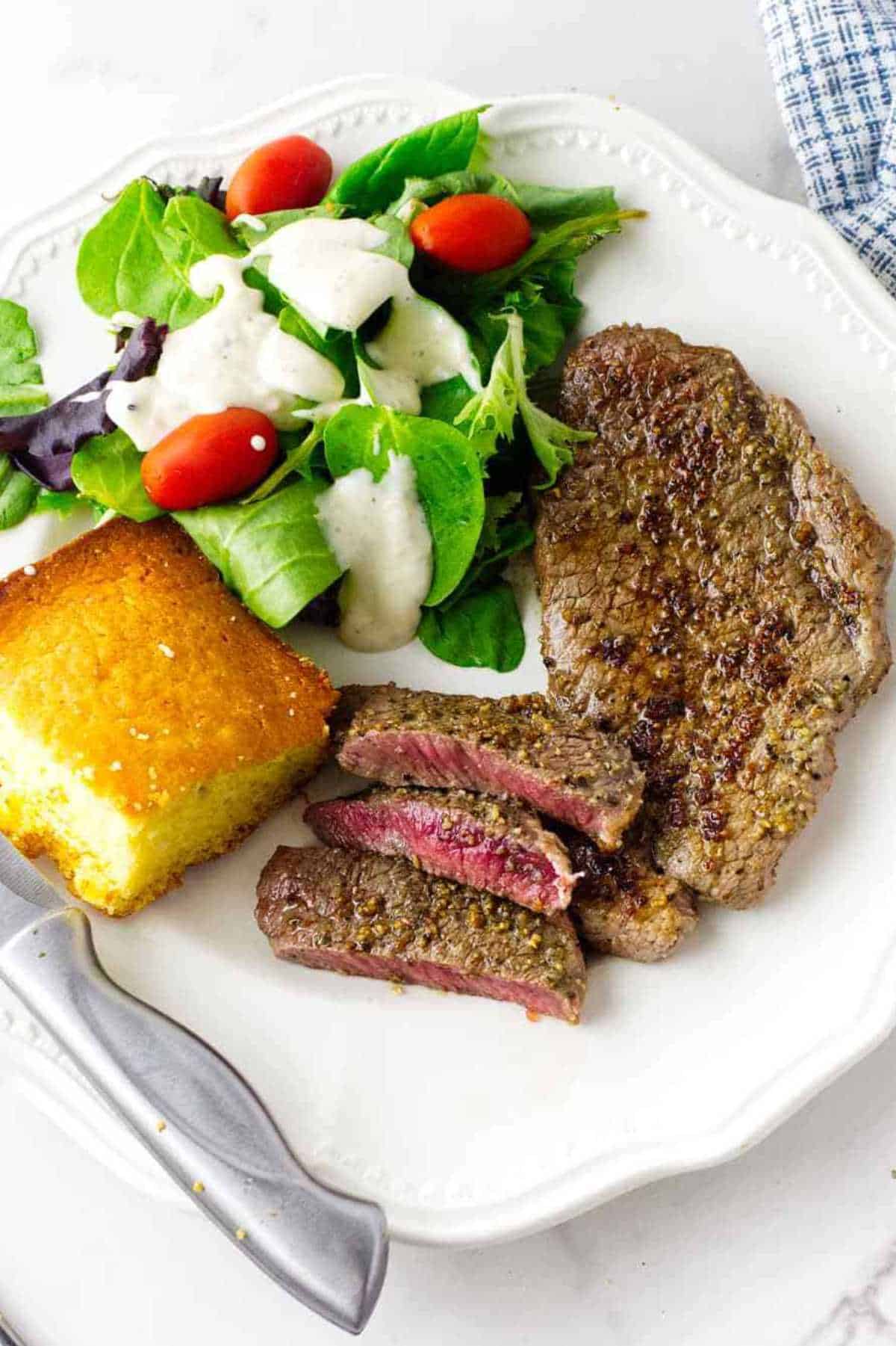 A plate with a grilled Denver steak, sliced to show a pink center, a green salad with grape tomatoes and creamy dressing, and a square piece of cornbread. A fork and knife are beside the plate.