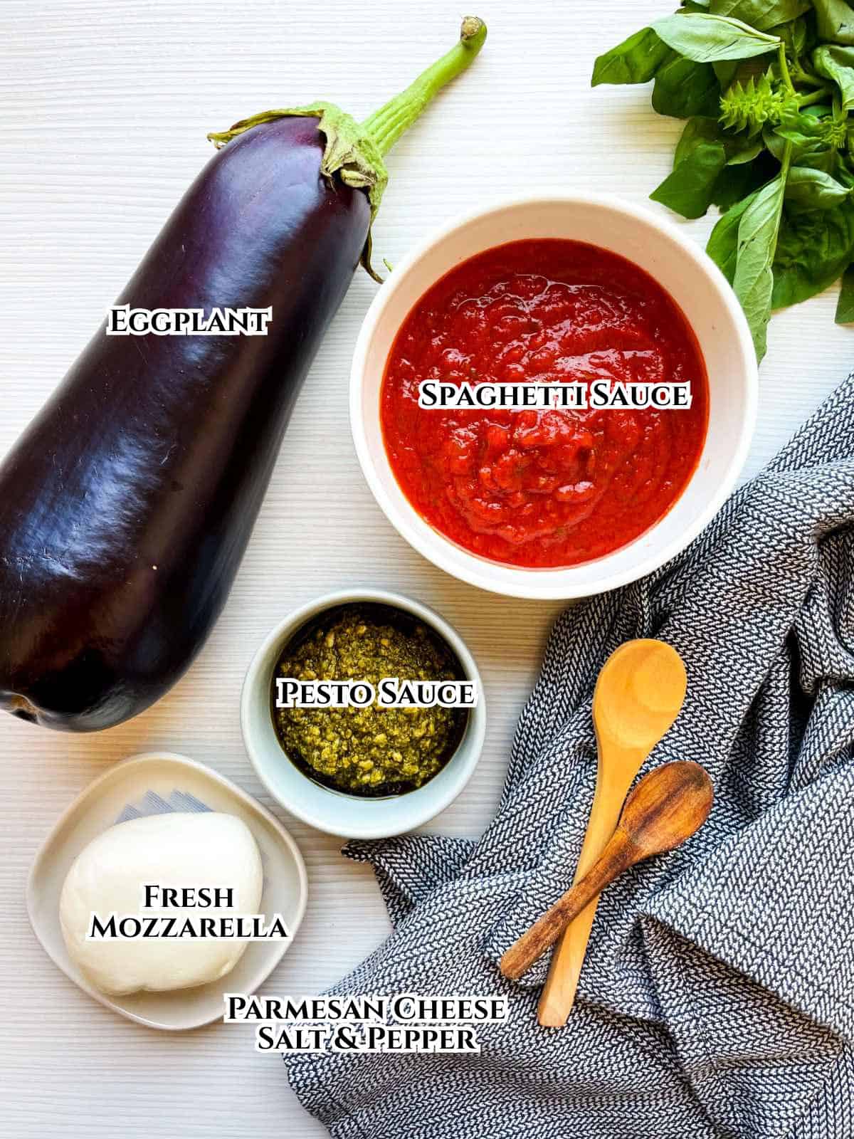 Flat lay of ingredients for an eggplant dish: a whole eggplant, a bowl of spaghetti sauce, a bowl of green seasonings, fresh mozzarella, basil leaves, and two wooden spoons on a gray towel. Labels identify each item.
