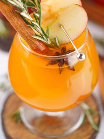 Two glasses of orange apple cider garnished with apple slices, cinnamon sticks, and rosemary sprigs. Red apples, cinnamon sticks, and a red patterned cloth are arranged in the background.